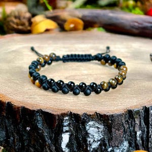 Op de afbeelding: Een zwart en goud kralenarmband. De armband heeft afwisselend zwarte en goudkleurige kralen, geregen op een zwart koord. De armband wordt gepresenteerd op een houten oppervlak.