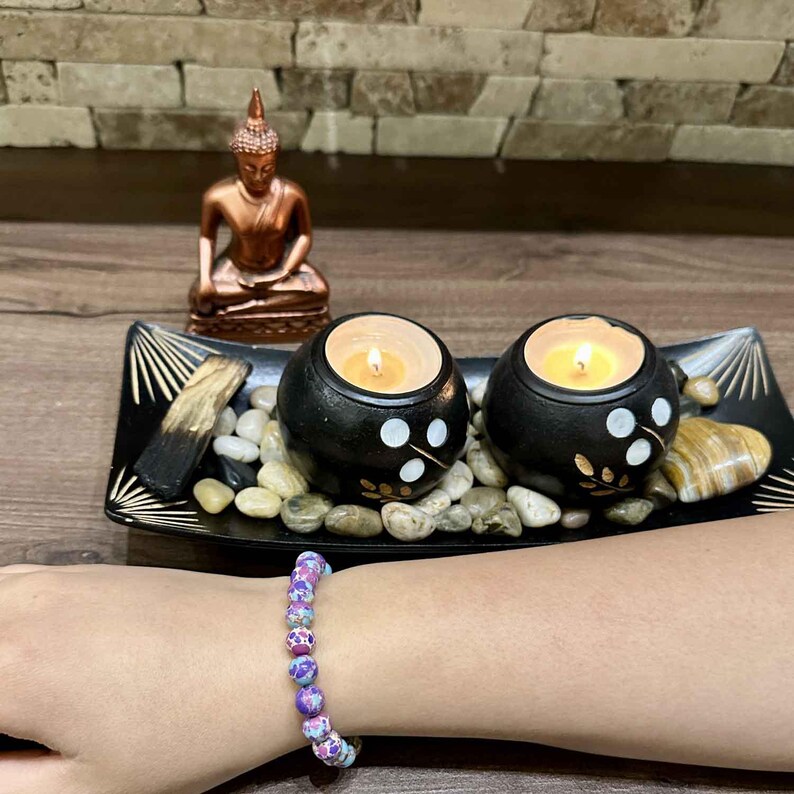 Pode incluir: Uma bandeja de madeira com duas velas pretas e brancas, uma pequena est&aacute;tua dourada de Buda e um bracelete de contas roxas e azuis.