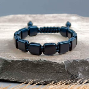 Può includere: Un braccialetto blu scuro con perline quadrate collegate da un cordoncino intrecciato. Il braccialetto è presentato su una superficie di pietra grigia strutturata. Il design è semplice ed elegante, adatto all'uso quotidiano. Il braccialetto misura circa 20 cm di lunghezza.