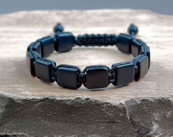 Puede incluir: Una pulsera azul oscuro con cuentas cuadradas unidas por un cordón trenzado. La pulsera se muestra sobre una superficie de piedra gris texturizada. El diseño es simple y elegante, adecuado para el uso diario. La pulsera mide unos 20 cm de largo.