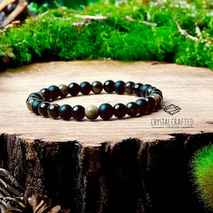 Op de afbeelding: Een armband van zwarte en gouden kralen met de tekst "CRYSTAL CRAFTED - Handmade" op een houten oppervlak.