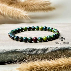 Pode incluir: Um bracelete de pedras preciosas verde, vermelho e amarelo sobre um fundo verde escuro. O bracelete é feito de pequenas contas redondas e é chamado de "Crystal Crafted Handmade".