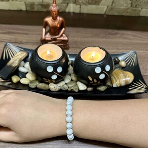 May include: White howlite beaded bracelet with a black and white candle holder with a lit candle in the center. The candle holder is surrounded by small stones and is on a black tray with gold accents.