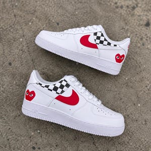 May include: A pair of white low-top sneakers featuring a red Nike swoosh, a black and white checkered pattern on the heel panel, and a red heart graphic with two black eyes positioned on the outer heel area of each shoe with white laces.