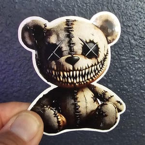 May include: A creepy, stitched-together teddy bear with a wide, toothy grin and X's for eyes. The bear is sitting on a white background.