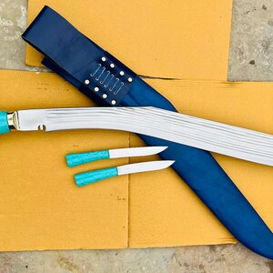 May include: A large, curved blade knife with a turquoise handle and two smaller knives, all with matching turquoise handles. The knife rests on a yellow surface with a blue sheath. The blade is silver and the sheath is dark blue.