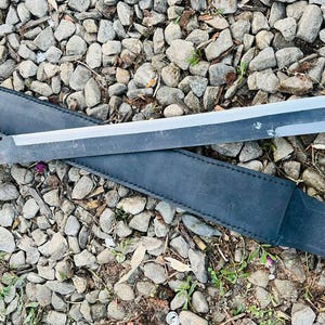 May include: A black leather scabbard with a silver blade sword. The sword has a black handle with a textured grip.