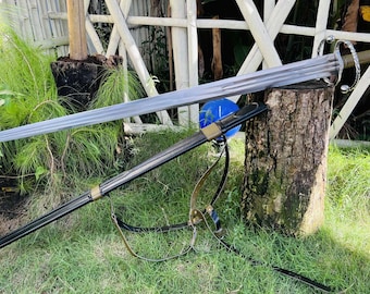 Puede incluir: Una espada larga y plateada con una hoja de doble filo descansa sobre un tocón de árbol. La espada tiene una empuñadura decorativa y se combina con una vaina negra con una correa de cuero. La escena está ambientada al aire libre con hierba verde y follaje.