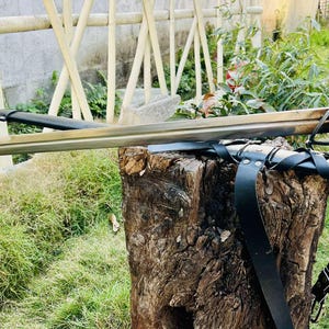 May include: A black sword with a metal guard and a leather strap. The sword is resting on a wooden stump with green grass in the background.