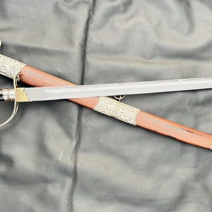 以下が含まれることがあります： 銀と黒の剣で、木製の柄と装飾的な銀のアクセントが付いています。剣は濃い灰色の表面に置かれています。