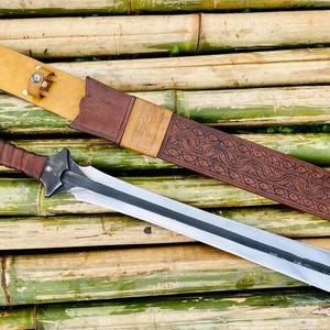 May include: A silver sword with a brown leather scabbard with intricate carvings. The scabbard has a gold-colored tip and a brown leather strap with metal accents.