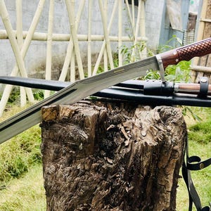 May include: A long, silver blade with a wooden handle and a black leather sheath. The blade is sharp and has a slight curve. The handle is decorated with a wood grain pattern. The sheath is attached to a black leather strap.