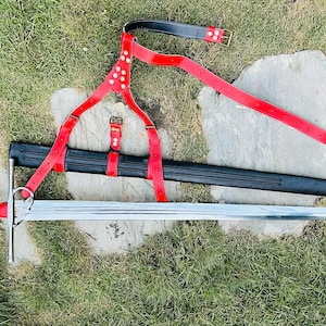 Puede incluir: Una espada de estilo medieval con una hoja plateada y un mango rojo, apoyada sobre hierba. También hay una vaina negra y un arnés de cuero rojo con cinturón. La longitud total de la espada es de aproximadamente 102 cm.
