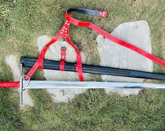 May include: A medieval-style sword with a silver blade and a red handle, resting on grass. A black scabbard and red leather harness with a belt are also present. The sword's total length is approximately 40 inches.