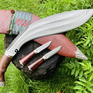 May include: A large, curved blade knife with a wooden handle and two smaller knives, all with wooden handles. The knives rest on a wooden surface, with a brown leather sheath. The sheath has decorative stitching and metal accents. The background is green.