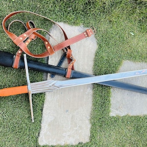 Puede incluir: Una espada plateada con una vaina de cuero marrón y una correa de cuero marrón. La espada está sobre una superficie de hierba verde.