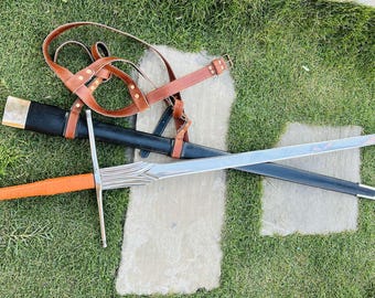 May include: A silver sword with a brown leather scabbard and a brown leather strap. The sword is lying on a green grassy surface.