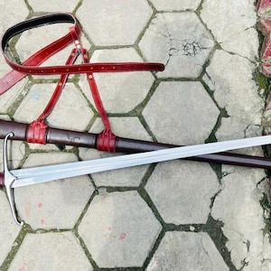 May include: A longsword with a silver-toned steel blade, a crossguard, and a red leather-wrapped handle with a dark metal pommel. The sword is accompanied by a dark brown leather scabbard and a red leather belt harness with metal buckles.