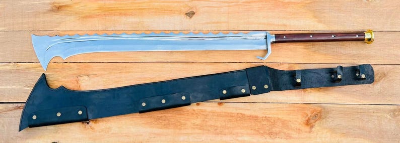 May include: A long, silver, serrated blade with a brown wooden handle and a gold accent. The blade is in a black leather scabbard with metal accents.