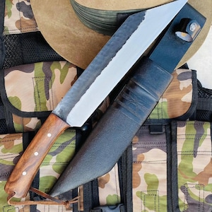 May include: A large,  handmade knife with a wooden handle and a black leather sheath. The knife has a  hammered finish and a sharp point. The knife is resting on a camouflage fabric background.