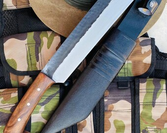 May include: A large,  handmade knife with a wooden handle and a black leather sheath. The knife has a  hammered finish and a sharp point. The knife is resting on a camouflage fabric background.