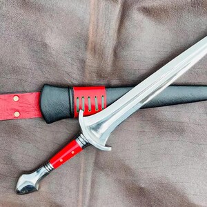 May include: A silver dagger with a red and black handle and a black leather sheath. The dagger is lying on a brown leather surface.