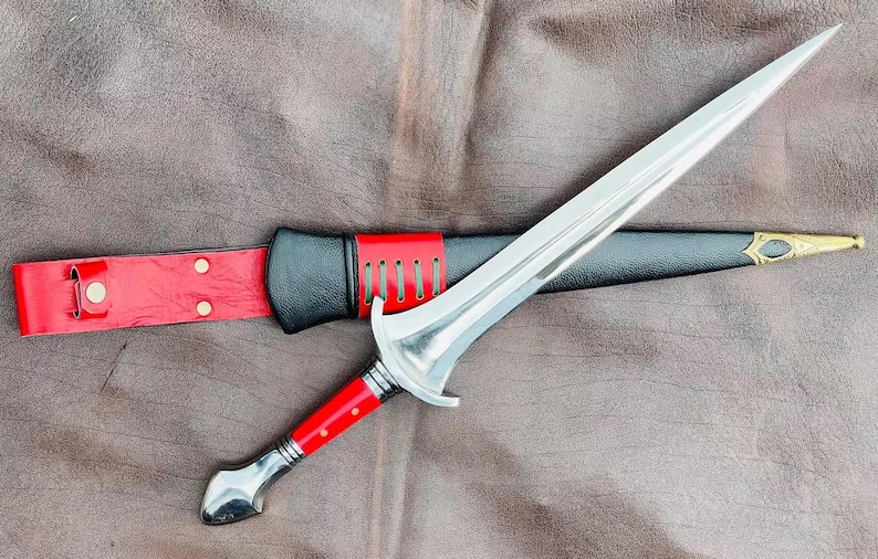 May include: A silver dagger with a red and black handle and a black leather sheath. The dagger is lying on a brown leather surface.