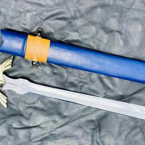 Puede incluir: Una espada plateada con empuñadura dorada y una vaina azul y marrón. La espada tiene un diseño de alas en la empuñadura.