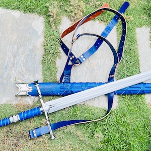 Puede incluir: Una espada larga y plateada con empuñadura y vaina azules, sobre hierba. La espada tiene una guarda y una correa de cuero. La vaina es azul con un patrón texturizado. El mango está envuelto en alambre azul y plateado.