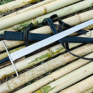 May include: A long, silver sword with a black handle and a black sheath, resting on a bed of bamboo stalks. The sword has a crossguard and a leather strap. The sheath has a gold-colored buckle.