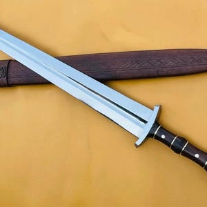 May include: A silver and black sword with a brown handle and sheath. The blade is long and sharp, with a crossguard. The handle is dark brown with silver accents. The sheath is brown and has a decorative pattern.