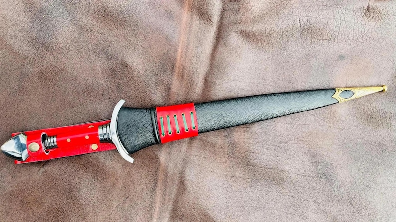May include: A black leather-wrapped dagger with a red leather sheath and a gold-colored tip. The dagger has a silver-colored metal guard and a red leather strap with five silver-colored rivets.