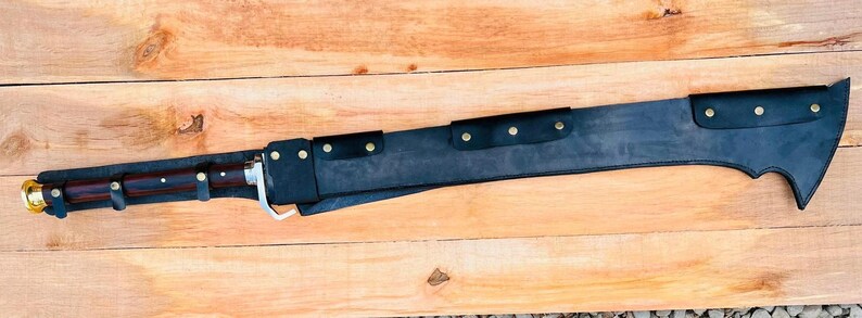 May include: A black and silver sword with a leather sheath and a wooden handle. The sword has a curved blade and a decorative design on the handle.