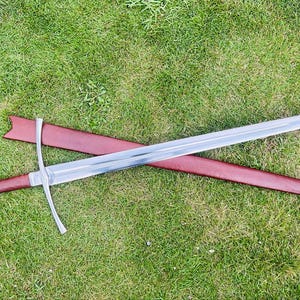 May include: A long, silver sword with a cross-shaped hilt and a brown handle lies on a red leather sheath. The sword is placed on green grass, highlighting its length and design.
