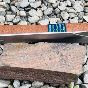 以下が含まれることがあります： 茶色の木製のハンドルと青い革製の鞘が付いた、大きな銀色のナイフ。ナイフは木の板の上に置かれています