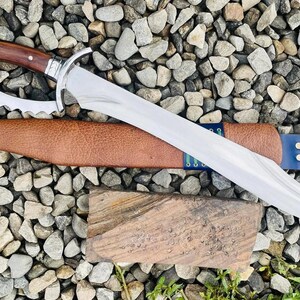 以下が含まれることがあります： 装飾的なハンドルと鋸歯状のエッジを持つ銀と茶色の剣。剣は青いストラップが付いた茶色の革製の鞘の上に置かれています。