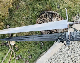 May include: A silver sword with a black leather scabbard. The sword has a long, straight blade and a crossguard. The scabbard has a strap and buckle.