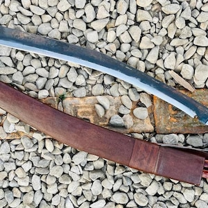 May include: A curved, silver-colored blade with a brown wooden handle and a brown leather sheath. The blade is resting on a bed of small, gray stones.