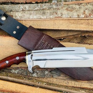 May include: A large, silver-colored knife with a brown wooden handle and a leather sheath. The knife has a sharp point and a curved blade.