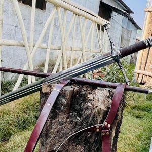 Könnte beinhalten: Ein silbernes Schwert mit einem braunen Ledergriff und einer Scheide. Das Schwert liegt auf einem Baumstumpf mit einem braunen Lederriemen.