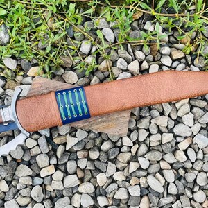 以下が含まれることがあります： 青と緑のパターンが施された青いストラップが付いた茶色の革製の鞘。鞘には、波状の刃を持つ銀色の曲がった刃が収められています。
