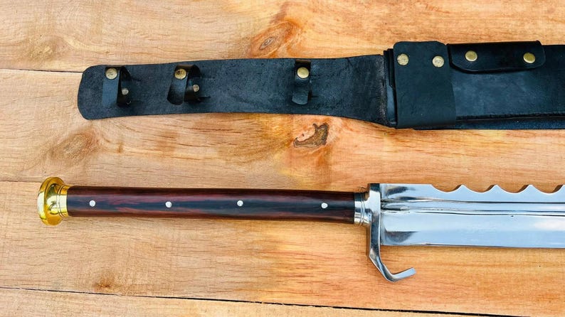 May include: A silver sword with a wooden handle and a black leather sheath. The sword has a serrated blade and a hook at the end.