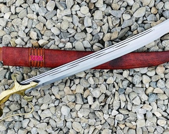 May include: A silver-colored sword with a decorative brass handle and a red leather scabbard. The sword has a curved blade with intricate details and is lying on a bed of small gray stones.