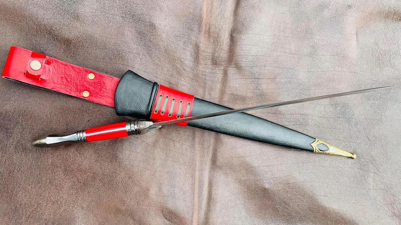May include: A black and red leather sheath with a silver-colored dagger with a black handle. The dagger is lying on a red leather surface.