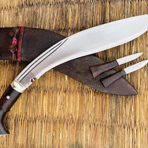 May include: A silver-coloured curved knife with a brown wooden handle and a leather sheath. The knife has a decorative pattern on the blade and the sheath has a red and brown design. Two smaller knives with brown wooden handles are also included.