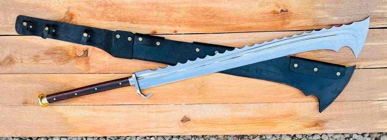 May include: A large, silver, serrated sword with a dark brown wooden handle and a black leather scabbard. The sword is lying on a wooden surface.