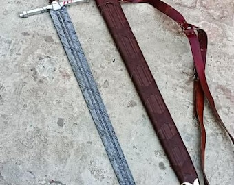 Puede incluir: Una espada larga con una hoja estampada, una guarda plateada y un mango marrón con detalles dorados. La espada está junto a una vaina de cuero marrón con punta dorada y una correa burdeos.