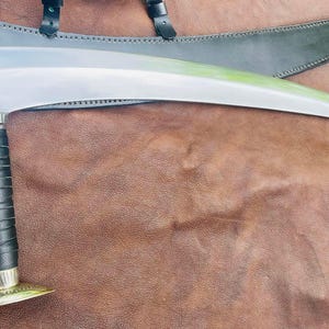 Puede incluir: Una hoja curva de color plateado con un mango de cuero negro y una empuñadura plateada. La hoja está apoyada sobre una superficie de cuero marrón.