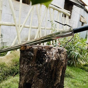 May include: A silver sword with a black leather handle resting on a tree stump. The sword is sharp and has a pointed tip. The sword is in a natural outdoor setting.