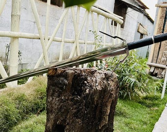 May include: A silver sword with a black leather handle resting on a tree stump. The sword is sharp and has a pointed tip. The sword is in a natural outdoor setting.
